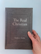 A hand holding a book titled 'The Real Christian: A Commentary on 2 Corinthians' by Gordon J. Keddie.