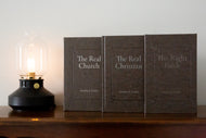 Three books titled 'The Real Church', 'The Real Christian', and 'The Right Path' on a wooden surface with a lamp in the background.